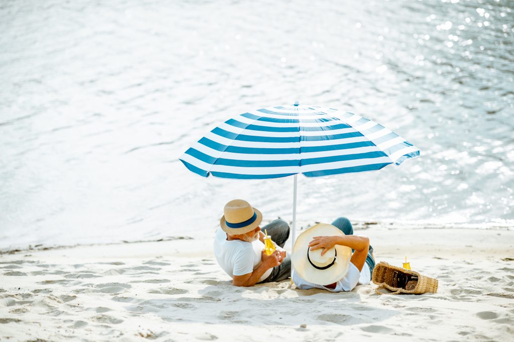 An edlerly couple sit on the beach in the shade of an umbrella and hats
