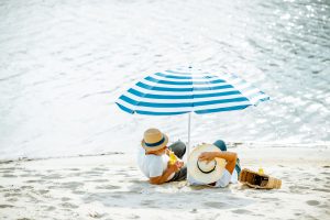 An edlerly couple sit on the beach in the shade of an umbrella and hats