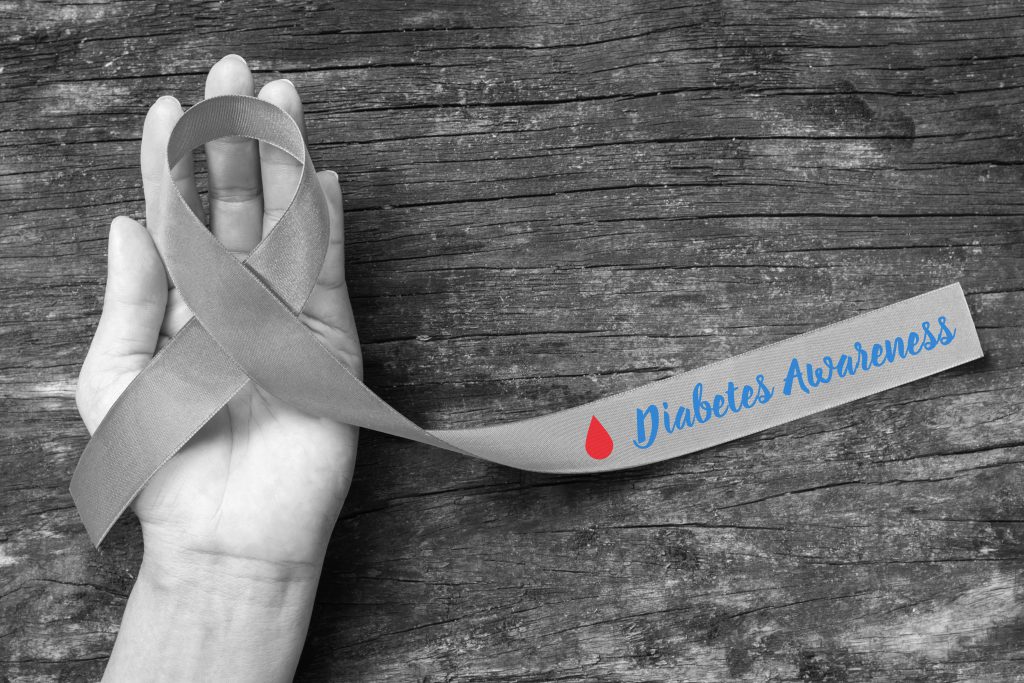 Black and white background with someone's hand holding a grey ribbon with the text "Diabetes Awareness" in blue and a red blood droplet