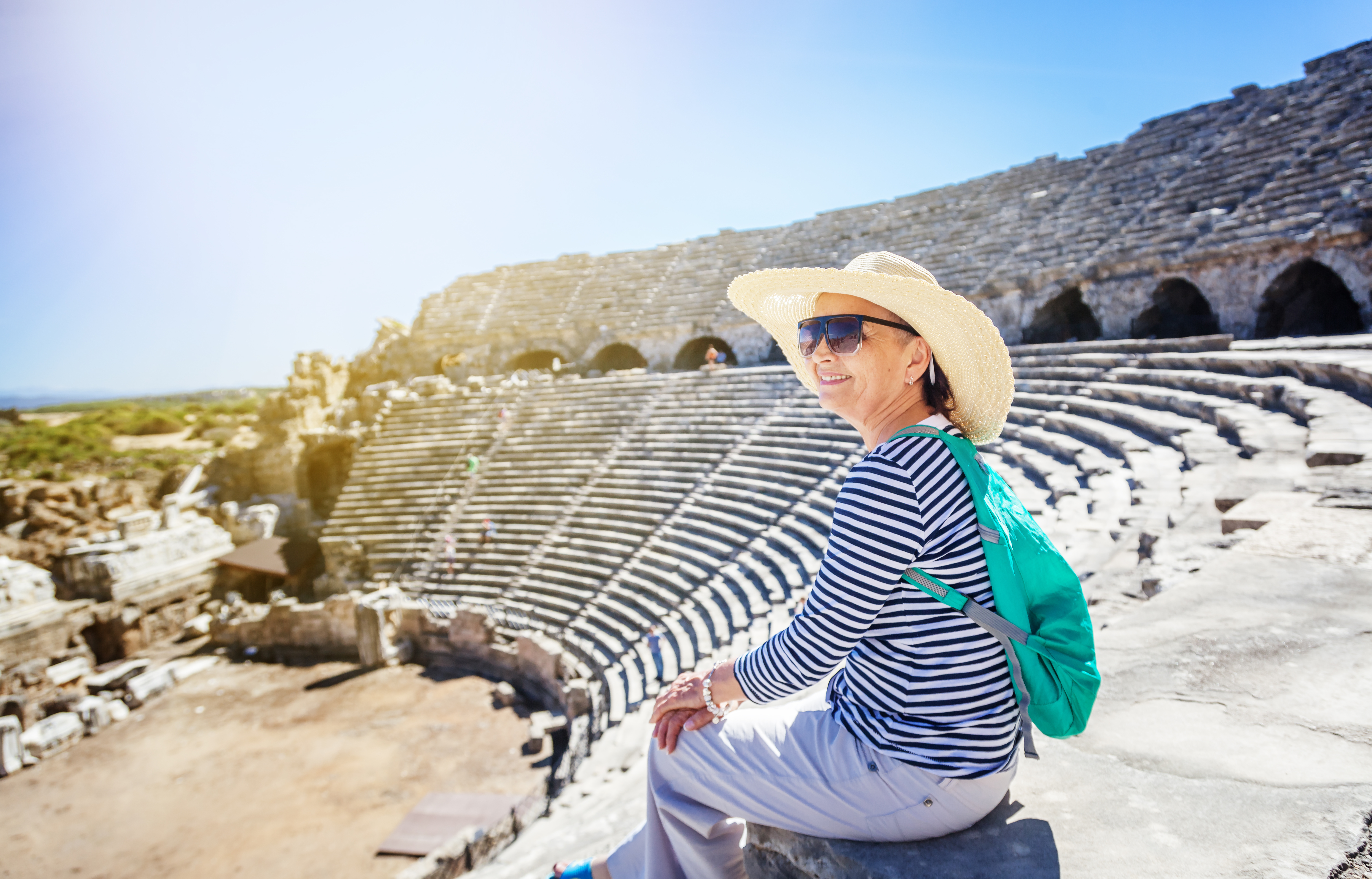 Elderly woman wears sunglasses and floppy hat as she sits smiling and admiring the scenery of an ancient amphitheater on a sunny day